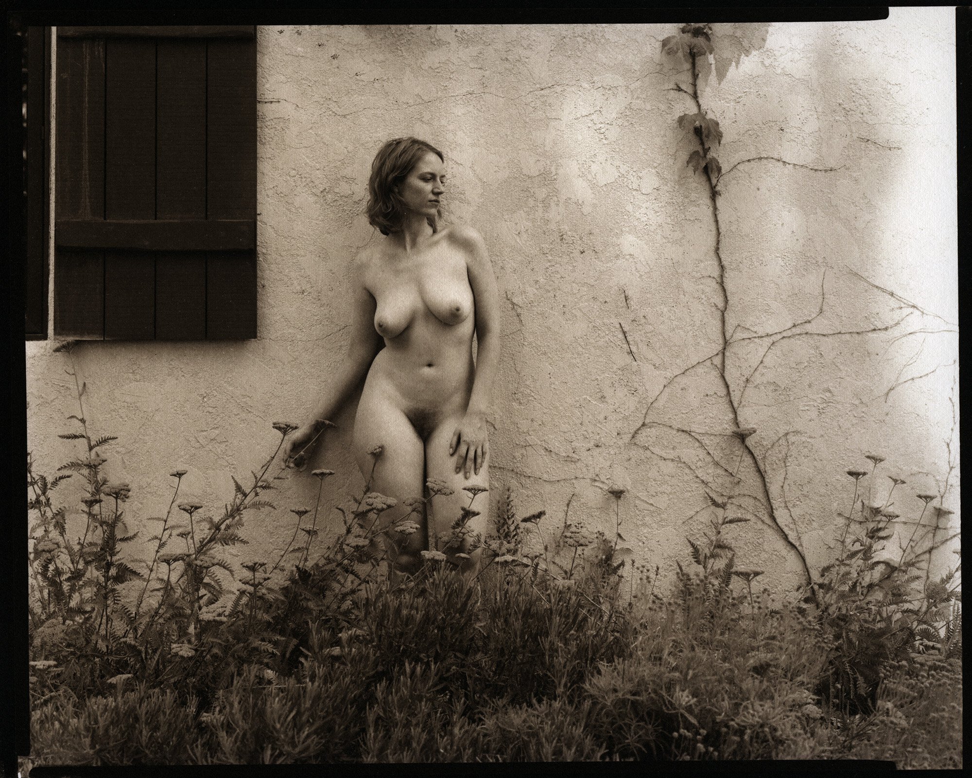 Sepia foto van een naakte vrouw tegen de muur van een oud huis met onkruid op de voorgrond